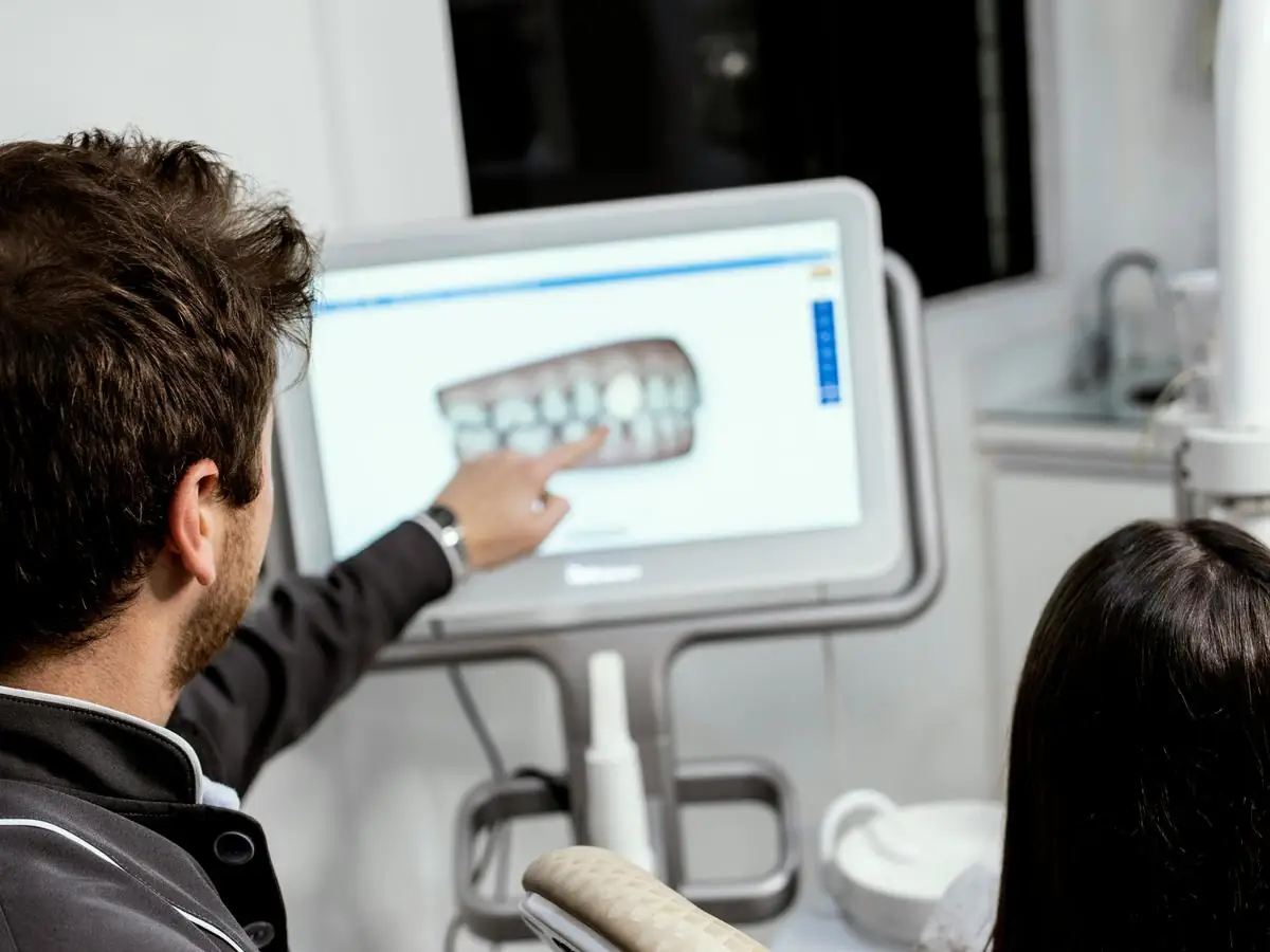 A dentist pointing at a screen showing a 3D scan of a patient’s teeth, explaining it to the patient sitting beside him.