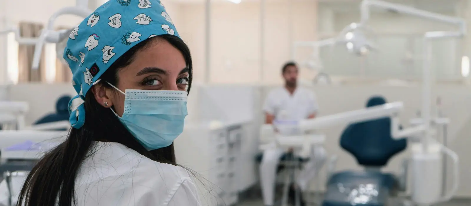 A dental assistant in the treatment room, turning towards the camera, with a colleague working at a chair behind her.